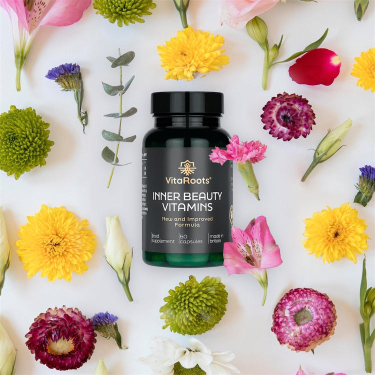 A black VitaRoots® “Inner Beauty Vitamins” bottle with a gold logo and label sits centered on a white background, surrounded by neatly arranged colourful flowers in pink, yellow, green, and purple, creating a clean, elegant flat-lay product scene.